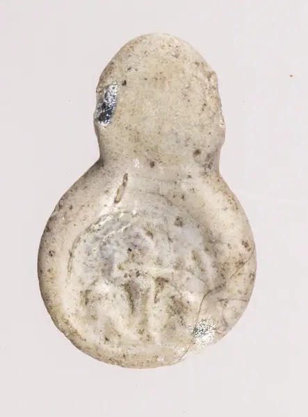 Amuletă de sticlă, de epocă romană târzie. Piesa este realizată din sticlă albă, cu partea principală, de formă circulară, și o prelungire destinată orificiului de agățare. Pe față a fost realizat un 